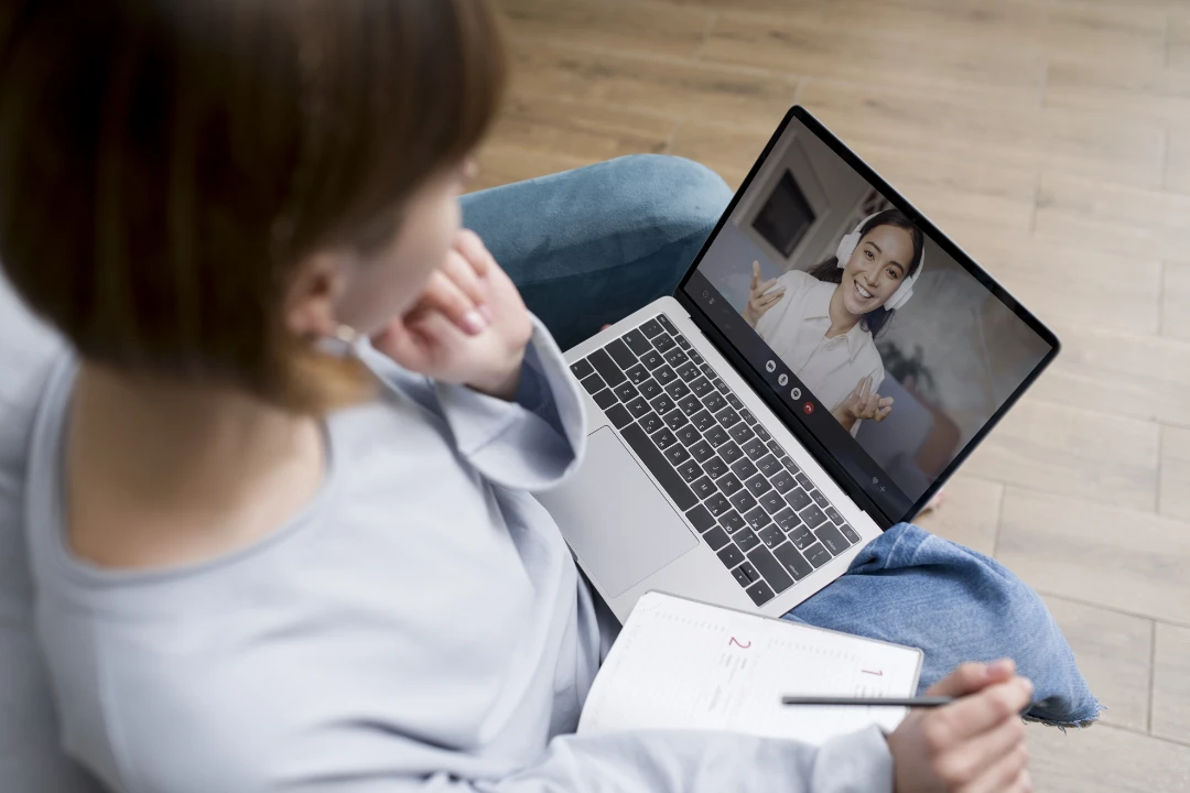 Persona sentada en un sofá tomando notas mientras realiza una videollamada en una computadora portátil, demostrando cómo atraer a tu paciente ideal con una web diseñada para psicólogos puede facilitar conexiones profesionales y efectivas en terapia online.