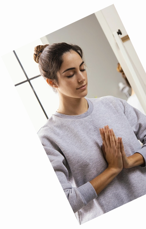 img-perfil-psic-7-c-der Una persona con los ojos cerrados y el pelo recogido en un moño, vestida con una sudadera gris, está de pie con las manos en posición de oración, creando una imagen serena perfecta para una página de destino compatible con SEO.