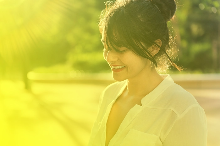 img-ter-Ind-2-e Una mujer con cabello oscuro recogido en un moño, vestida con un top blanco, sonríe mientras está parada afuera en un día soleado. Los rayos del sol y las sombras son visibles en el fondo, lo que agrega profundidad a este momento sereno que a menudo se captura para las páginas de destino.