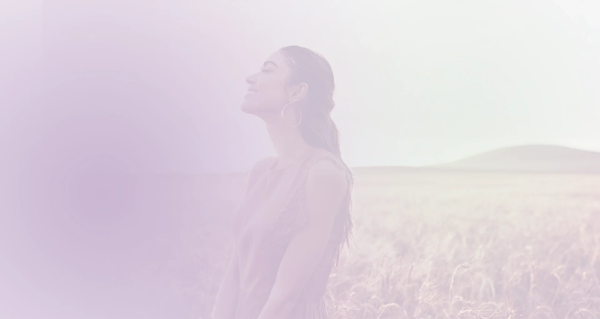 img-ter-ind-4a2 Una mujer de pelo largo está parada en un campo de hierba alta, mirando hacia arriba y sonriendo. La imagen, perfecta para la página de inicio de un sitio web, tiene un tinte púrpura suave.