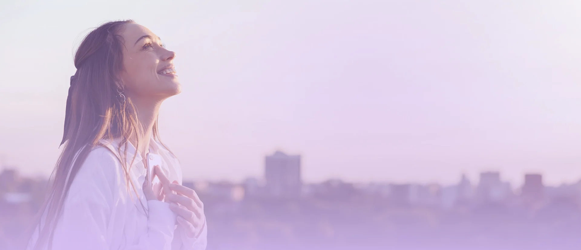 img-ter-ind-5a Una persona con el pelo largo se encuentra al aire libre, mirando hacia arriba y sonriendo con el horizonte de una ciudad al fondo. La imagen, perfecta para landing pages o estrategias SEO, tiene un tinte violeta.