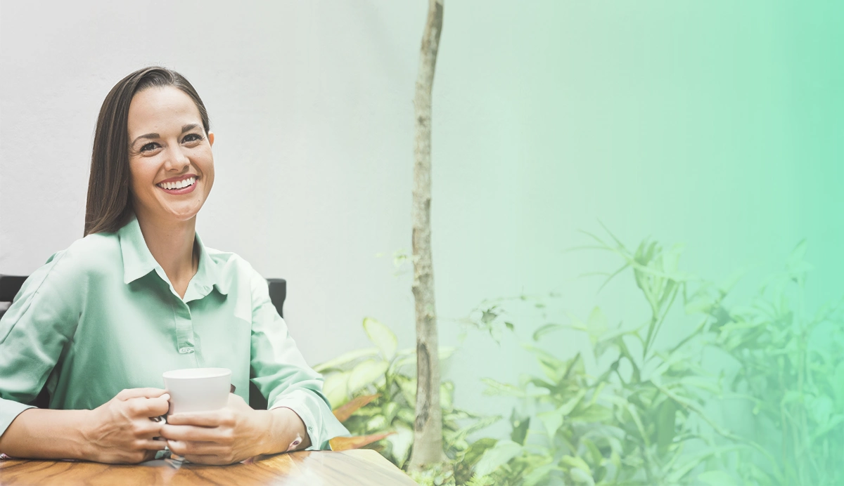 img-ter-ind-7c Una mujer con una camisa verde claro está sentada en una mesa de madera al aire libre, sosteniendo una taza blanca y sonriendo a la cámara. Se ve un fondo de jardín borroso en el lado derecho de la imagen, lo que crea una atmósfera acogedora perfecta para cualquier optimización de sitio web o imagen de página de destino.