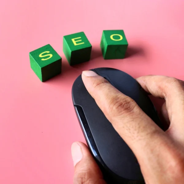 Una mano que sostiene un ratón de computadora negro junto a tres bloques verdes con letras amarillas que deletrean "SEO" sobre un fondo rosa, lo que simboliza cómo optimizar un sitio web para destacar en las búsquedas de Google.