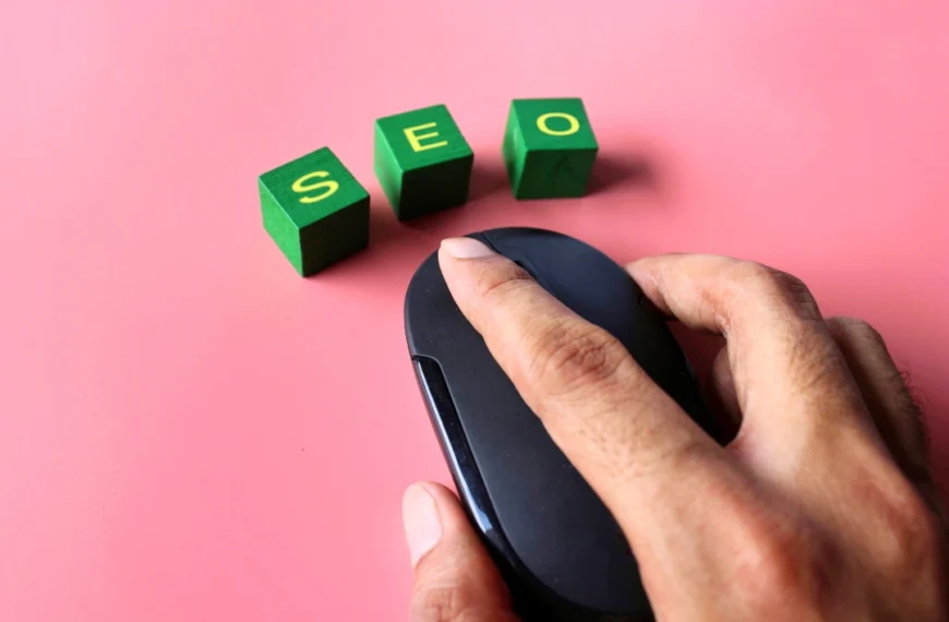 Una mano que sostiene un ratón de computadora negro junto a tres bloques verdes con letras amarillas que deletrean "SEO" sobre un fondo rosa, lo que simboliza cómo optimizar un sitio web para destacar en las búsquedas de Google.