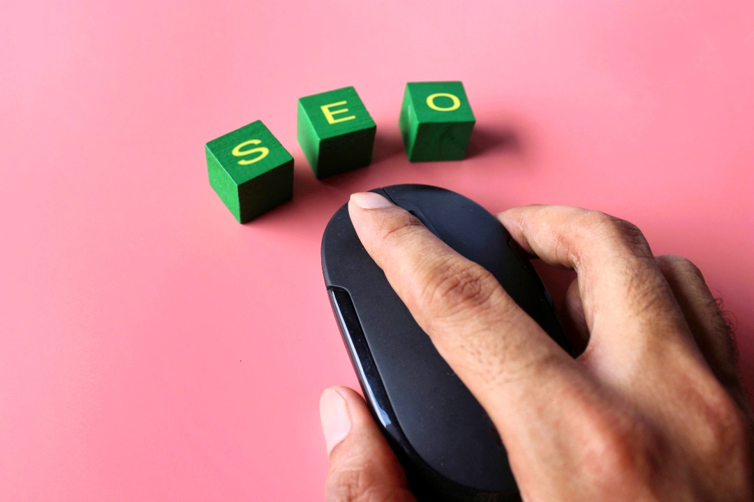 Una mano que sostiene un ratón de computadora negro junto a tres bloques verdes con letras amarillas que deletrean "SEO" sobre un fondo rosa, lo que simboliza cómo optimizar un sitio web para destacar en las búsquedas de Google.